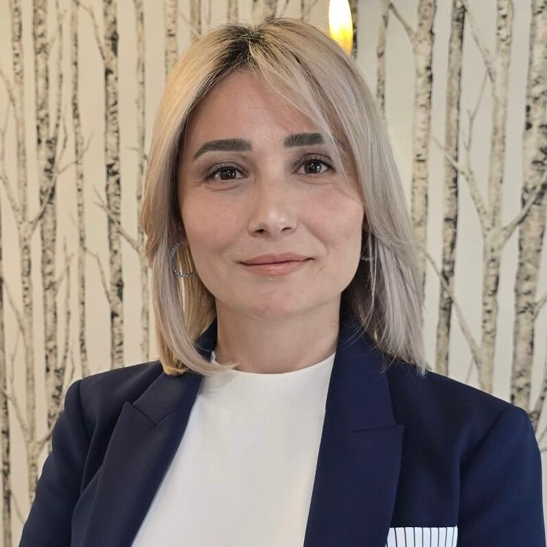 Professional headshot of a blonde woman with shoulder-length hair, wearing a white shirt and a dark blazer.